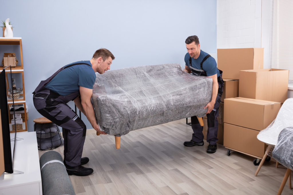 Portrait Of Two Young Happy Male Movers Carrying Wrapped Sofa In New House